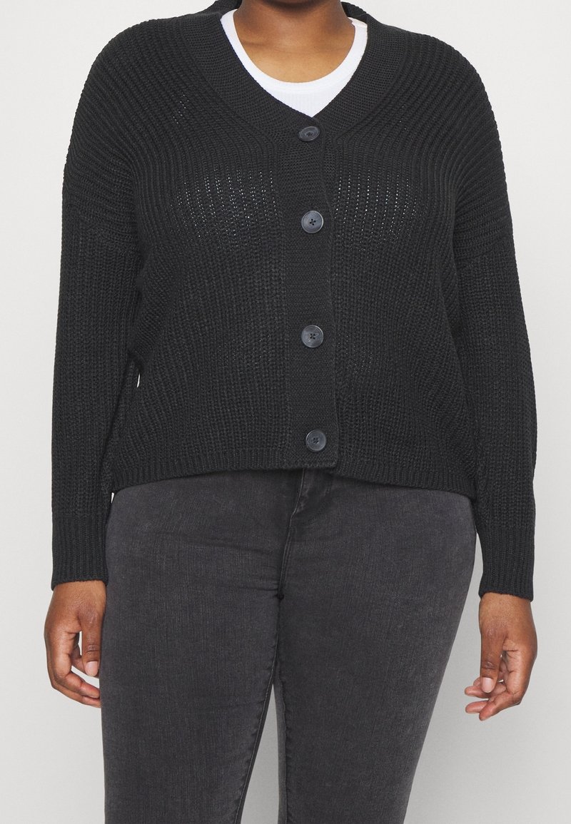 Femme portant un cardigan en tricot noir boutonné sur une chemise blanche et un pantalon gris foncé, visible du cou aux hanches.