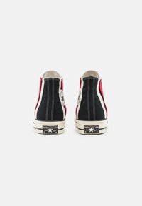 Scarpe sneakers alte in tela nere e rosse con cuciture bianche, suola in gomma e toppe con logo circolare sui lati. Materiale testurizzato, design classico.