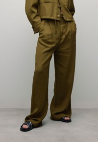 Pantalons en tissu mélangé lin, taille haute, de couleur vert olive, avec des poches latérales et une taille à cordon, associés à des sandales à glissière noires avec clous.