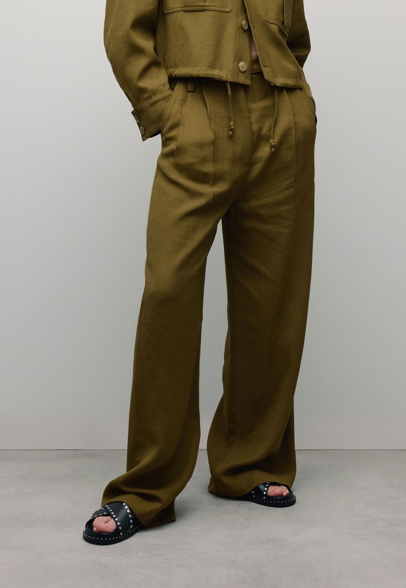 Pantalons en tissu mélangé lin, taille haute, de couleur vert olive, avec des poches latérales et une taille à cordon, associés à des sandales à glissière noires avec clous.