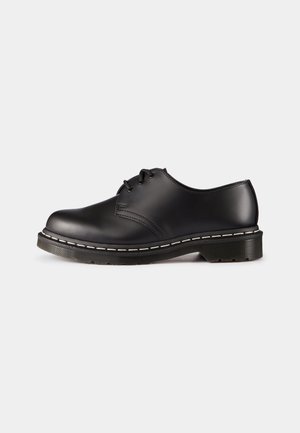 Chaussure basse en cuir noir à lacets avec semelle épaisse et détails de couture blanche sur fond clair uni.