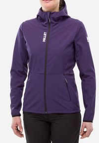Femme portant une veste softshell violette avec fermeture éclair à l'avant, capuche, poches zippées et logo "MILLET" blanc sur la poitrine et la manche.
