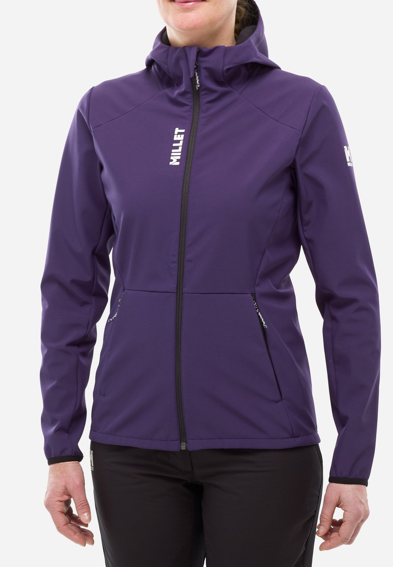 Femme portant une veste softshell violette avec fermeture éclair à l'avant, capuche, poches zippées et logo "MILLET" blanc sur la poitrine et la manche.