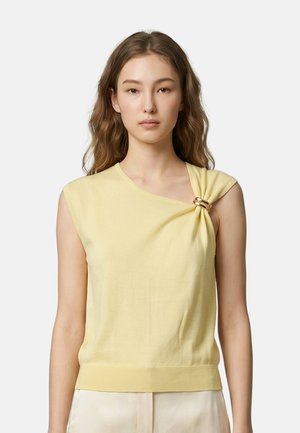 Jonge vrouw met lang golvend haar die een mouwloze lichtgele top draagt met een gouden ringdetail op één schouder.