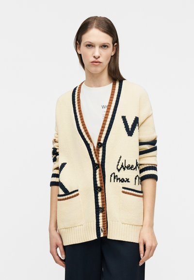 Femme portant un cardigan crème oversize avec des rayures noires et marron, boutonné à l'avant, avec des poches avant et des détails de texte noirs sur les manches et le corps.