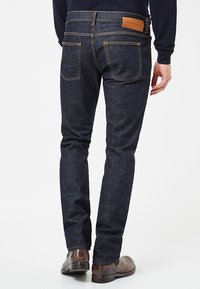 Vaqueros de mezclilla azul oscuro con un corte slim, que presentan costuras en amarillo en contraste, dos bolsillos traseros y una etiqueta de cuero marrón en la cinturilla.
