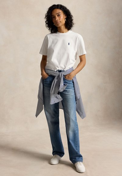 Kobieta o kręconych włosach ubrana w białą koszulkę, niebieskie dżinsy, białe tenisówki i koszulę w paski zawiązaną w talii, stoi z rękami w kieszeniach.