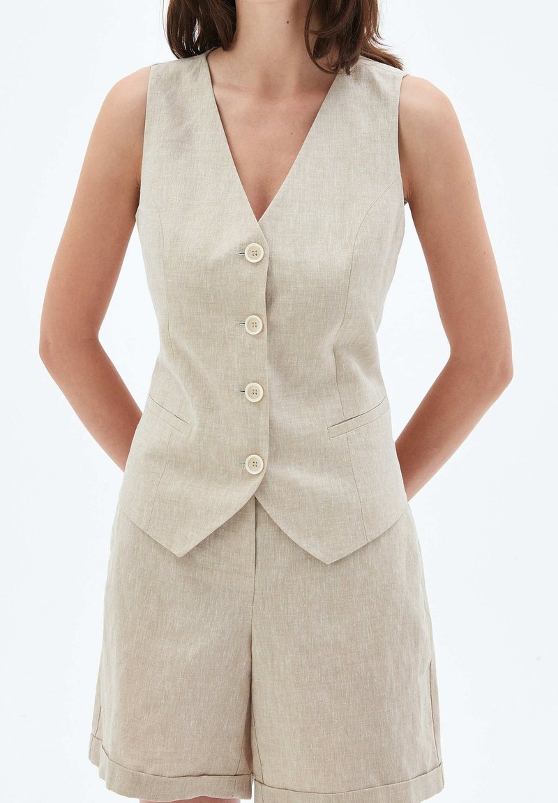 Beige linneväst med djup V-ringning, fem knappar framtill och två sidfickor. Tyget har ett strukturerat, lätt och luftigt utseende.