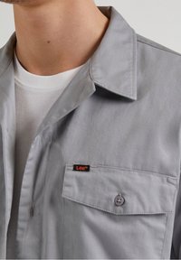 Maglietta grigia a maniche corte realizzata in tessuto misto cotone, con colletto, taschino con bottone e un piccolo logo Lee nero e arancione.