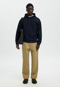 Sudadera azul marina con capucha y cordones, combinada con pantalones de pierna ancha beige. El modelo lleva un pequeño bolso de hombro gris. Un calzado oscuro completa el look.