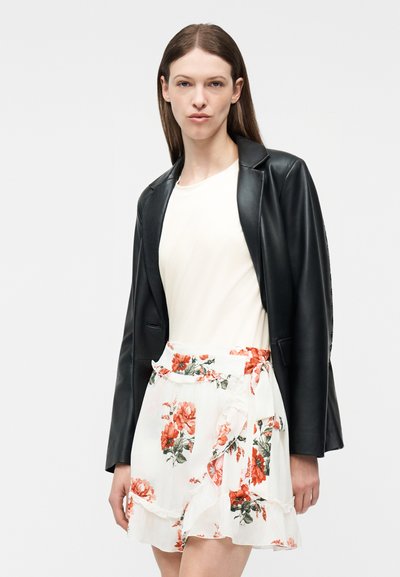 Γυναίκα που φορά μαύρο δερμάτινο μπουφάν πάνω από λευκό τοπ και λευκή φλοράλ φούστα τύπου wrap με κόκκινες και πράσινες λεπτομέρειες, στέκεται μπροστά σε ένα απλό φόντο.