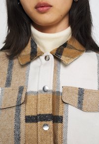 Närbild av en person som bär en beige, svart och vit rutig jacka med silverfärgade tryckknappar över en krämfärgad polotröja.