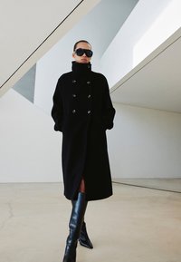 Czarny płaszcz z mieszanki wełny z wysokim kołnierzem, zapinany na dwa rzędy guzików, z przednimi kieszeniami i bocznymi rozcięciami, noszony z czarnymi kozakiami do kolan i okularami przeciwsłonecznymi.