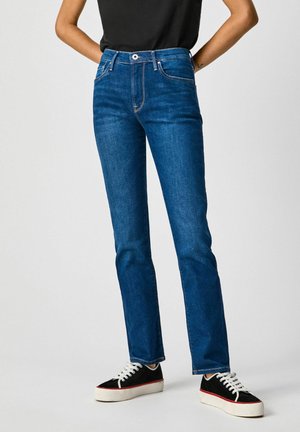 Persoon draagt straight-leg blauwe jeans met vijfzakkenontwerp, gecombineerd met zwart-witte lage sneakers, staat met handen achter de rug.