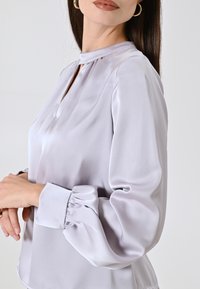 Blusa in seta di un leggero grigio chiaro con colletto alto e scollatura a chiave. Presenta maniche a sbuffo con polsini arricciati e una vestibilità comoda.