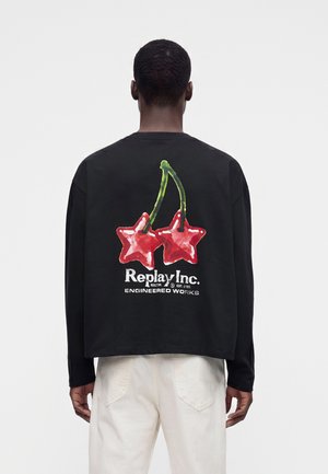 Person trägt ein schwarzes Langarmshirt mit roten sternförmigen Kirschen und weißem "Replay Inc." Text auf dem Rücken, kombiniert mit weißen Hosen.