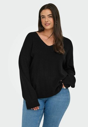 Femme aux longs cheveux bruns portant un pull noir à col en V ample et un jean bleu clair, se tenant avec une main dans la poche.