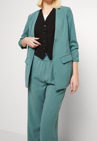 Teal blazer met zakken over een zwart vest met vijf knopen, gecombineerd met bijpassende tapered trousers, wat zorgt voor een gecoördineerde look.