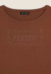 Camiseta de algodón marrón con escote redondo y un logo decorativo de piedras de strass "FREDDY" contorneado con tachuelas. Textura de tela suave.