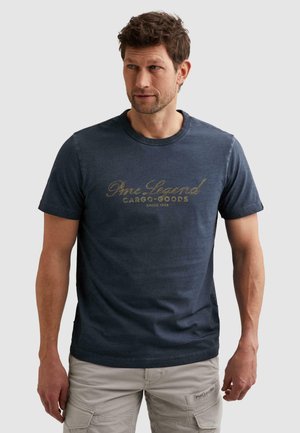 Man draagt een donkerblauw PME Legend Cargo Goods T-shirt en lichtgrijze cargoshorts, staand tegen een effen lichtgrijze achtergrond.