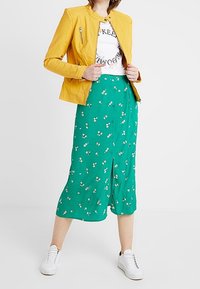 Giacca gialla in similpelle, t-shirt bianca con grafica, gonna midi a stampa floreale verde e sneakers bianche, con dettaglio di spacco davanti.