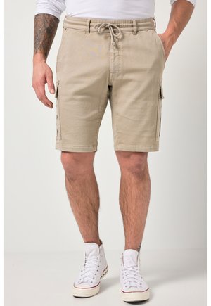 Beige cargoshorts med sidelommer båret af en stående mand med tatoveret arm, kombineret med hvide højtstående sneakers.