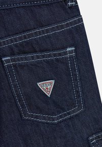 Τσέπη από σκουρομπλε τζιν με λευκή ραφή και μια μικρή τριγωνική ετικέτα Guess ραμμένη στο κέντρο της τσέπης.
