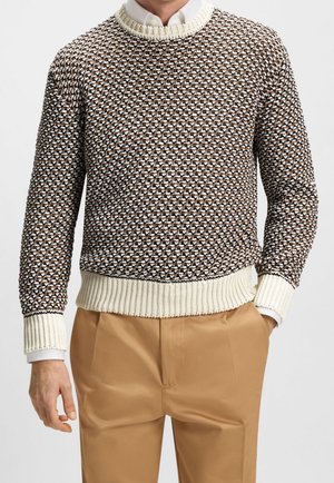 Mann trägt braunen, gemusterten Strickpullover mit cremefarbenen Rippbündchen an Ärmeln und Saum, weißes Hemd mit Kragen und beige Hose, eine Hand in der Tasche.