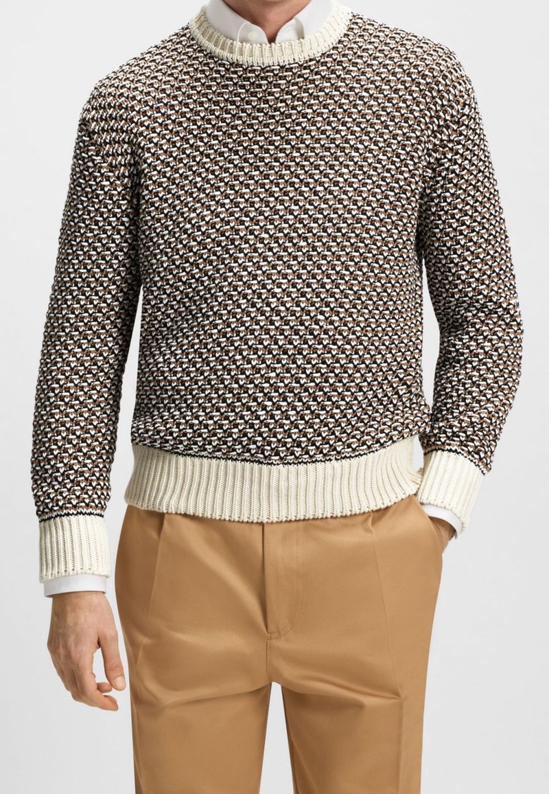 Homme portant un pull en tricot marron à motifs avec des poignets et un bas côtelés crème, une chemise blanche à col et un pantalon beige avec une main dans la poche.