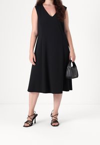 Černé šaty bez rukávů s výstřihem do V, rozšířeným tvarem a délkou do kolen. Oblečené s černými sand ály na vysokém podpatku a držící malou černou kabelku.