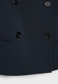 Tmavě námořnický dvouřadý kabát s čtyřmi černými knoflíky a dvěma horizontálními průstřihovými kapsami, orámovaný nařasenými švy.