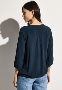 Marineblauwe, lichte blouse met driekwart mouwen, een ronde hals en geplooide details op de achterkant, gecombineerd met lichte jeans.