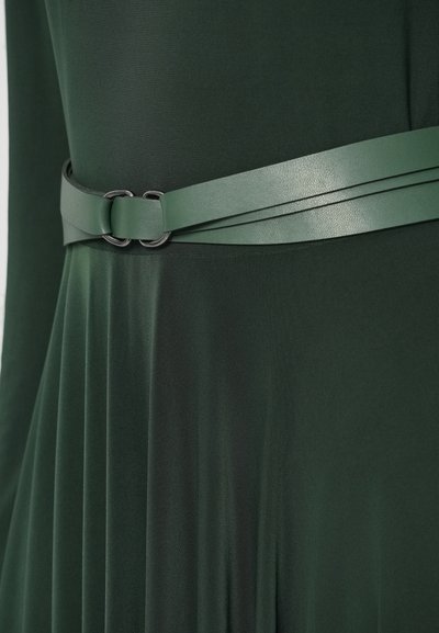 Zaļas kleitas ar plīsētu svārku un platām matētām zaļām jostām ar melnas metāla sprādzi slēgšanai, izceļot gludu auduma faktūru.