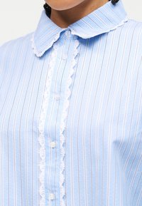 Κοντινό πλάνο ενός ατόμου που φοράει μπλε και λευκό ριγέ μπλούζα με δαντέλα στολισμένη κατά μήκος της σειράς κουμπιών και του γιακά.