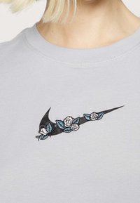 Ljusgrå sweatshirt med en svart Nike-logotyp prydd med vita rosor och blåa blad broderade längs designen.