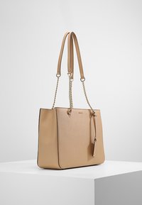 Sac fourre-tout en cuir beige avec deux anses, détail en chaîne dorée et tirette de fermeture éclair. Texture lisse avec des accents de quincaillerie minimalistes.