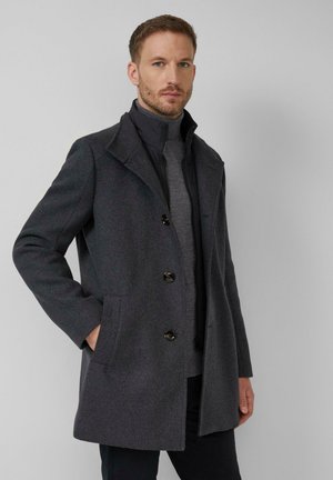 Mężczyzna ubrany w ciemnoszary wełniany płaszcz na czarnej kurtce na zamek i szary golf, stojący na tle jasnoszarego tła.