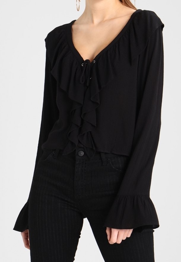 Blouse noire à manches longues avec un devant volanté, un décolleté en V et un détail à lacets. Dotée de manches évasées et d'un tissu texturé. Associée à un pantalon noir à rayures.