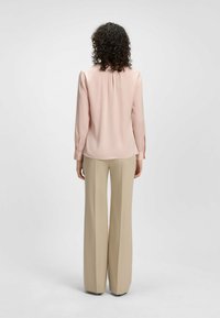 Blusa a maniche lunghe di colore rosa chiaro con scollo rotondo, texture liscia e dettaglio a pieghe sul retro, abbinata a pantaloni larghi beige.