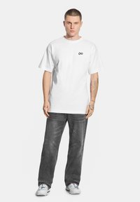 Biały bawełniany T-shirt z małym czarnym logo nieskończoności na piersi, zestawiony z luźnymi szarymi dżinsami i szarymi trampkami.