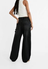 Czarne spodnie cargo z szerokimi nogawkami i wieloma kieszeniami, wykonane z denimu, o luźnym kroju i elastycznych mankietach. Widok z tyłu, pokazujący dopasowane szwy.