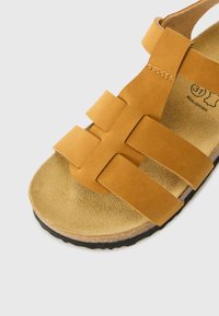 Sandale en cuir tan avec de larges brides présentant un design tissé. Semelle intérieure douce et texturée ainsi qu'une semelle extérieure en caoutchouc noir durable.