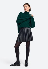 Grüner Strickpullover mit hohem Kragen, kombiniert mit einem schwarzen, plissierten Kunstlederrock, schwarzen blickdichten Strumpfhosen und glänzenden schwarzen Loafers.