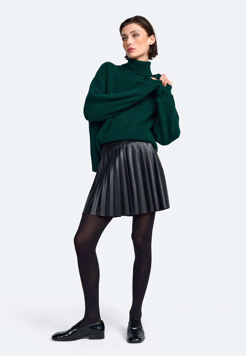 Grüner Strickpullover mit hohem Kragen, kombiniert mit einem schwarzen, plissierten Kunstlederrock, schwarzen blickdichten Strumpfhosen und glänzenden schwarzen Loafers.