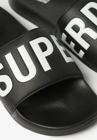Sorte slidesandaler med en bred rem, der har "SUPER" i fed hvide bogstaver. Overfladen har en glat struktur og afrundet form.