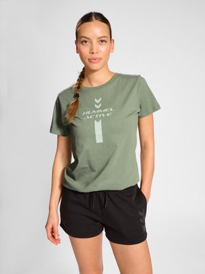 Donna con capelli castani intrecciati che indossa una maglietta verde "Hummel Active" e pantaloncini sportivi neri, in piedi con una mano in tasca.