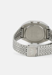 Montre métallique argentée avec un boîtier carré, un bracelet texturé, deux boutons sur le côté et un logo gravé à l'arrière.