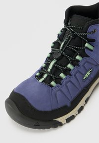 Zapato de senderismo morado que presenta una combinación de materiales de ante y malla, detalles en negro, suela texturizada y un sistema de cordones integrado para mayor soporte.