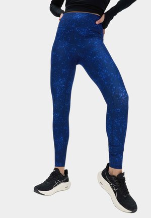 Osoba ubrana w niebieskie legginsy w wzory i czarne buty do biegania, ręce spoczywają na biodrach na jednolitym tle.