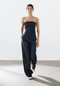Denim-Top ohne Träger mit tailliertem Oberteil, kombiniert mit weiten, hoch geschnittenen Hosen. Beides in einem tiefen Indigo-Farbton mit glatter Textur.